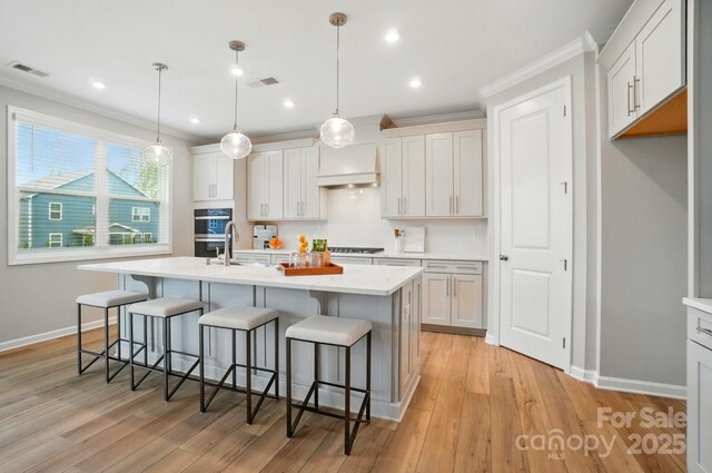 9930 Cask Way, Huntersville, NC 28078 - kitchen with custom range hood, a center island with sink, a kitchen bar, double oven, and light wood-style flooring