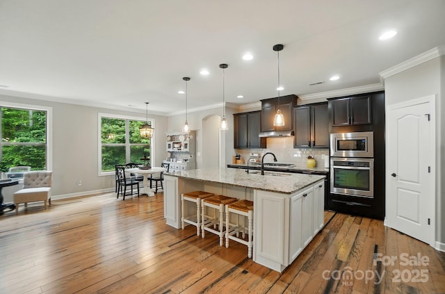 110 St Sophia Ct, Mooresville, NC 28117 - kitchen featuring appliances with stainless steel finishes, an island with sink, decorative backsplash, light stone countertops, and crown molding