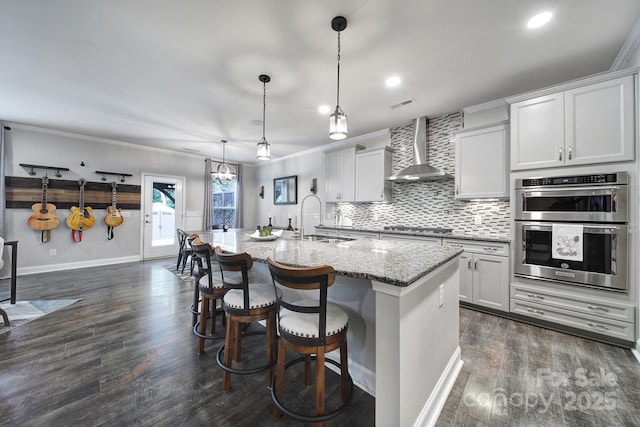 5424 Tilley Manor Dr, Matthews, NC 28105 - kitchen featuring wall chimney exhaust hood, double oven, light stone counters, an island with sink, and ornamental molding