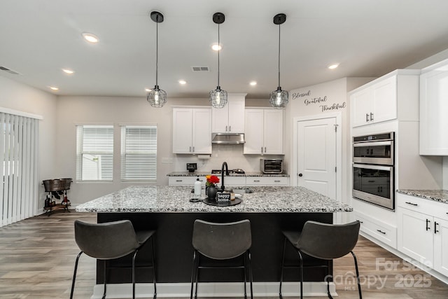 9274 Dunwich Dr, Gastonia, NC 28056 - kitchen with under cabinet range hood, double oven, a sink, an island with sink, and wood finished floors