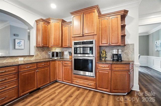 5916 Four Wood Dr, Matthews, NC 28104 - kitchen with stainless steel double oven, dark wood-style floors, arched walkways, ornamental molding, and dark stone counters