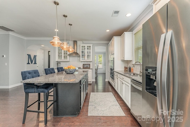 2606 Cheverny Pl, Concord, NC 28027 - kitchen featuring appliances with stainless steel finishes, wall chimney exhaust hood, a kitchen island, arched walkways, and white cabinets