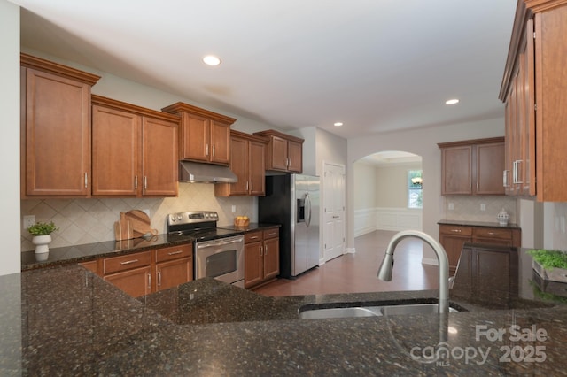 11922 Kimberfield Rd, Matthews, NC 28105 - kitchen featuring stainless steel appliances, under cabinet range hood, arched walkways, brown cabinetry, and dark stone countertops