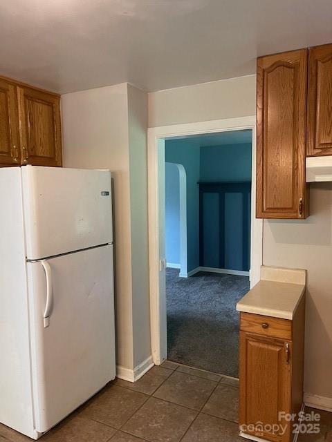 kitchen with freestanding refrigerator, under cabinet range hood, brown cabinets, dark tile patterned flooring, and dark carpet