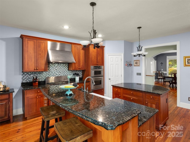 303 S Braeside Ct, Asheville, NC 28803 - kitchen with wall chimney exhaust hood, an island with sink, double oven, light wood-style floors, and arched walkways