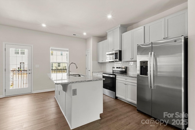 1204 May Apple Dr, Matthews, NC 28104 - kitchen featuring appliances with stainless steel finishes, a kitchen island with sink, decorative backsplash, light stone counters, and dark wood-type flooring
