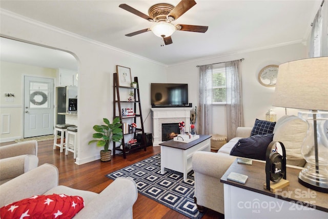 1219 Flowe Dr, Matthews, NC 28104 - living room with crown molding, a ceiling fan, dark wood-style floors, a warm lit fireplace, and arched walkways