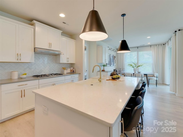 16108 Langston Dr, Charlotte, NC 28278 - kitchen featuring under cabinet range hood, light wood finished floors, decorative backsplash, white cabinets, and a kitchen island with sink