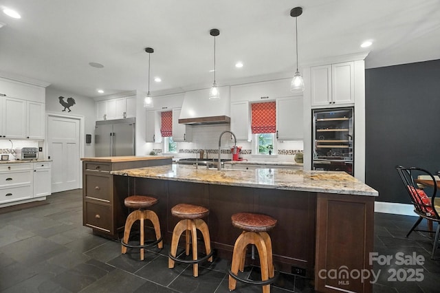 5005 Craftsman Ridge Dr, Matthews, NC 28104 - kitchen with stainless steel built in fridge, beverage cooler, a kitchen island with sink, white cabinetry, and custom range hood