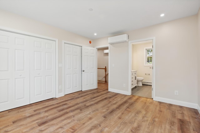 unfurnished bedroom with baseboard heating, baseboards, an AC wall unit, light wood-style floors, and multiple closets