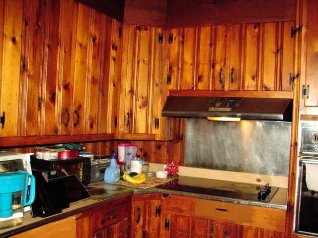 kitchen featuring oven, wood walls, black electric cooktop, and wall chimney range hood