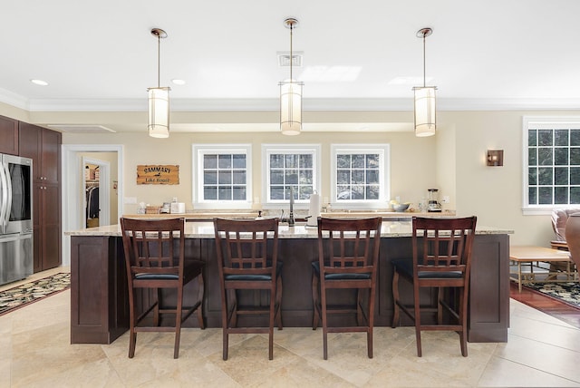 kitchen with light stone counters, stainless steel fridge, decorative light fixtures, and a center island with sink