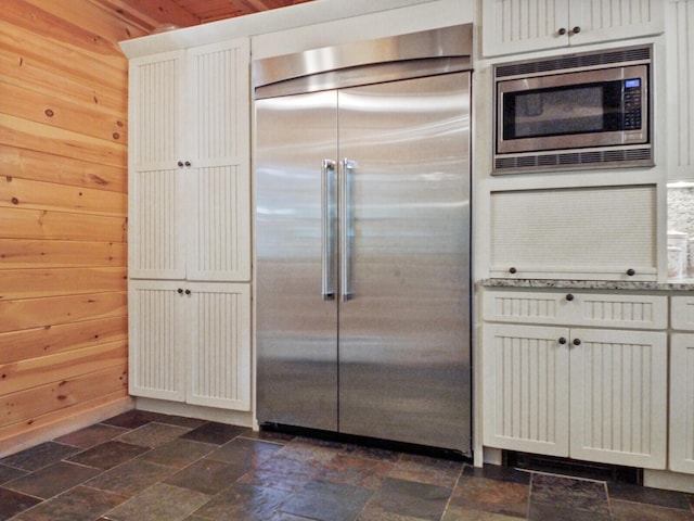 kitchen featuring built in appliances, stone finish flooring, white cabinetry, and wooden walls