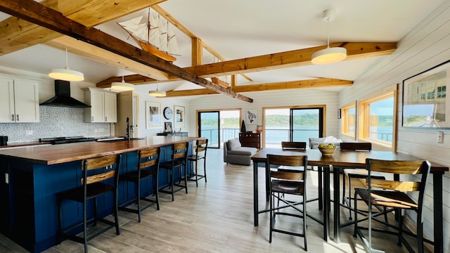 kitchen with a breakfast bar, decorative light fixtures, white cabinetry, wooden counters, and wall chimney range hood