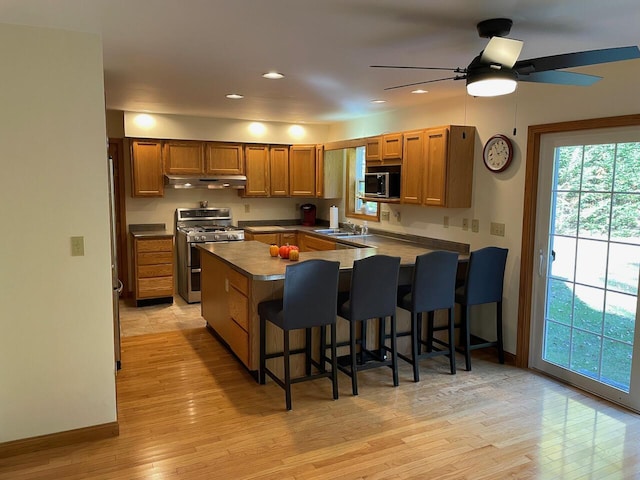 kitchen featuring a peninsula, light wood-style flooring, brown cabinetry, under cabinet range hood, and appliances with stainless steel finishes