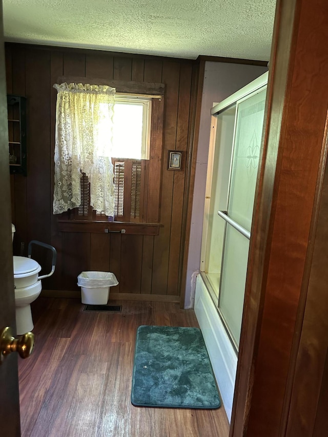 bathroom featuring toilet, wood walls, hardwood / wood-style flooring, a textured ceiling, and shower / bath combination with glass door