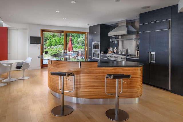 kitchen featuring appliances with stainless steel finishes, light hardwood / wood-style flooring, a breakfast bar area, and wall chimney range hood