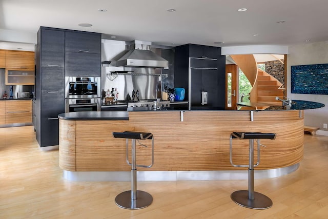 kitchen with light hardwood / wood-style flooring, double oven, a kitchen breakfast bar, and wall chimney exhaust hood