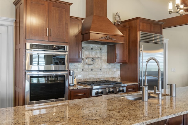 kitchen featuring appliances with stainless steel finishes, tasteful backsplash, hanging light fixtures, light stone counters, and custom range hood