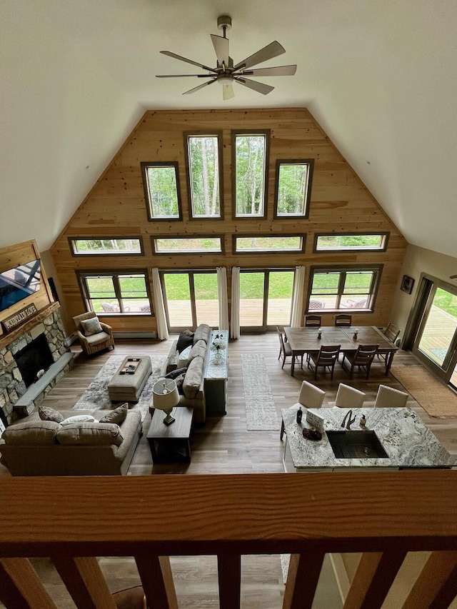 living room with high vaulted ceiling, wood walls, a stone fireplace, light wood-style flooring, and a ceiling fan