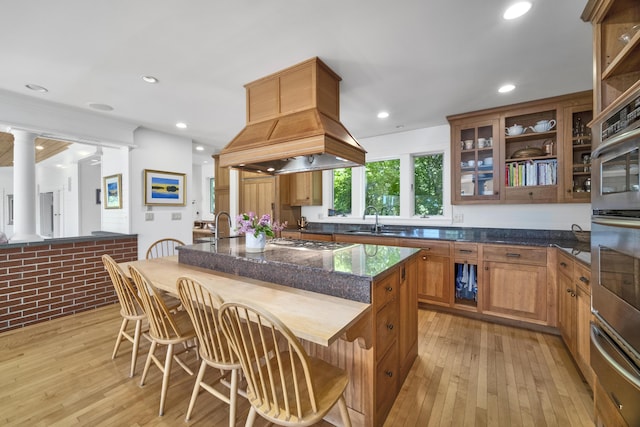 kitchen featuring custom exhaust hood, dark stone counters, sink, light hardwood / wood-style flooring, and an island with sink