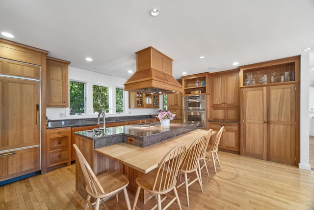 kitchen with premium range hood, dark stone counters, light hardwood / wood-style floors, a center island with sink, and appliances with stainless steel finishes