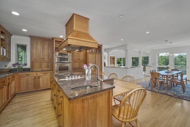 kitchen with sink, an island with sink, light hardwood / wood-style floors, decorative light fixtures, and appliances with stainless steel finishes