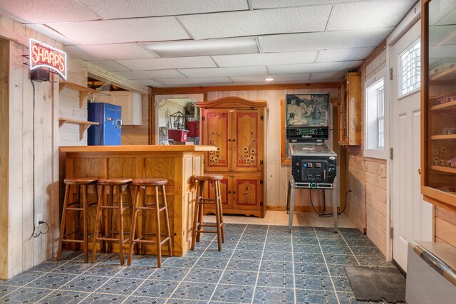 bar featuring wood walls, black refrigerator, tile floors, and a drop ceiling