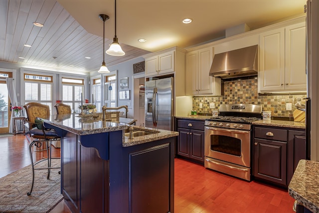 kitchen with light stone countertops, appliances with stainless steel finishes, wall chimney range hood, hanging light fixtures, and a kitchen island with sink