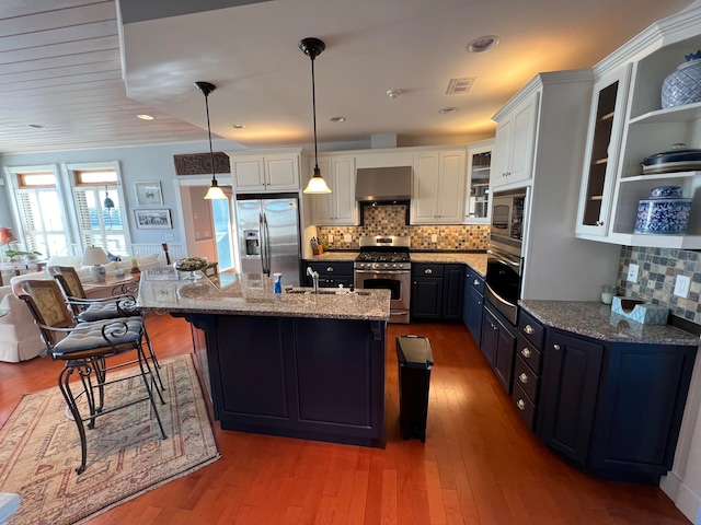 kitchen featuring a center island with sink, stainless steel appliances, pendant lighting, wall chimney exhaust hood, and white cabinets