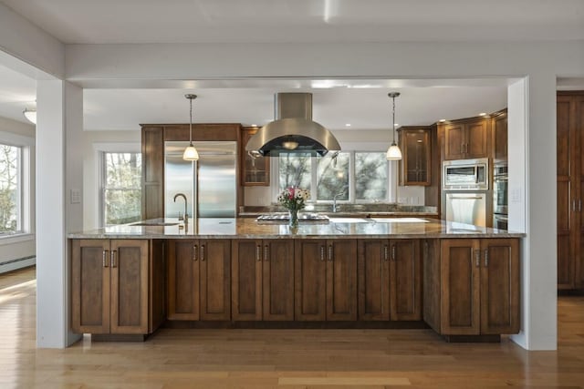 kitchen with island exhaust hood, pendant lighting, built in appliances, and dark stone counters