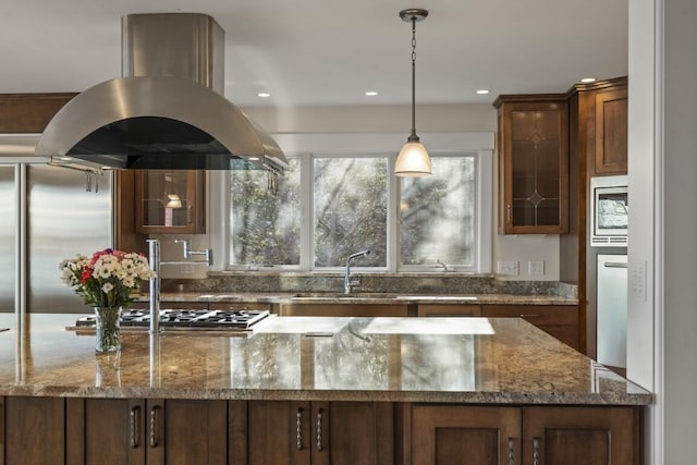 kitchen with island range hood, decorative light fixtures, sink, dark stone counters, and built in appliances