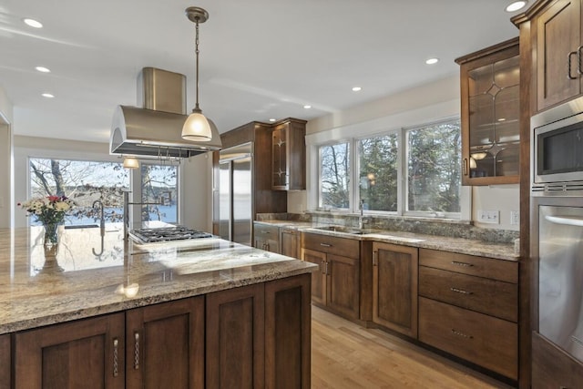 kitchen featuring island range hood, sink, hanging light fixtures, built in appliances, and light stone counters