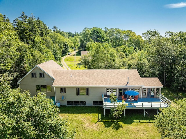 rear view of house featuring a forest view, a shingled roof, a lawn, and a wooden deck