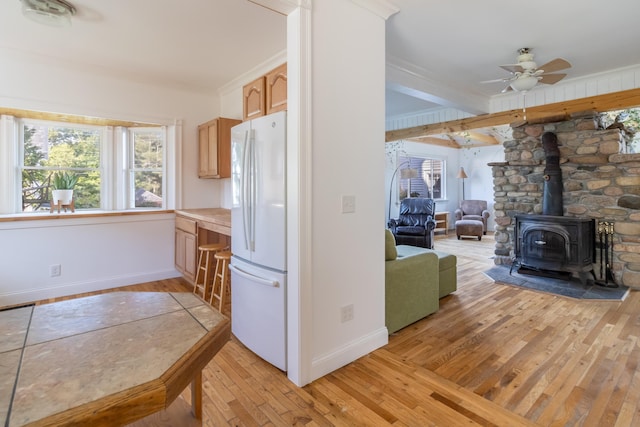 kitchen with light wood-style floors, white fridge with ice dispenser, light countertops, ceiling fan, and a wood stove