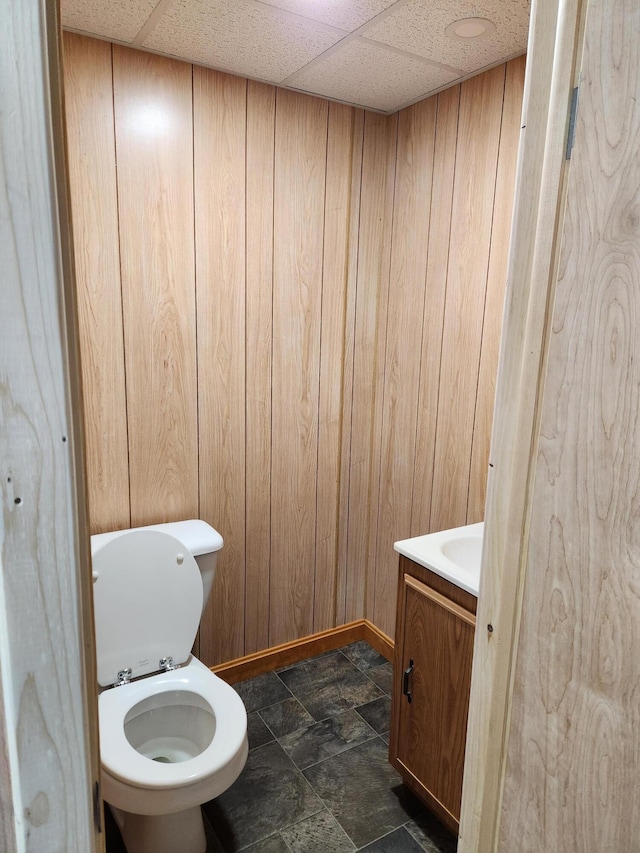 half bath featuring a drop ceiling, wood walls, vanity, and toilet