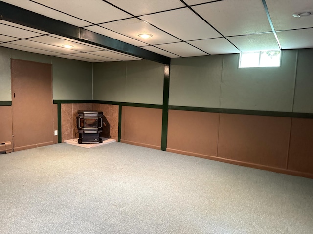 below grade area featuring carpet floors, a drop ceiling, a wood stove, and a decorative wall