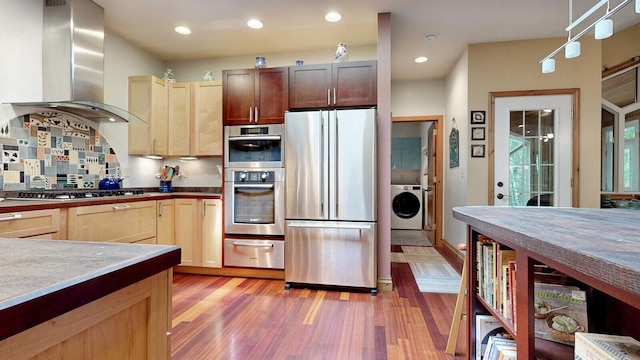 kitchen with a warming drawer, appliances with stainless steel finishes, wood finished floors, washer / dryer, and wall chimney exhaust hood