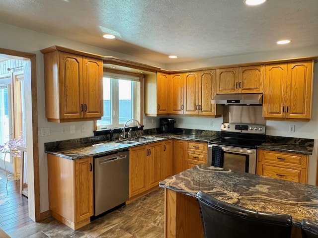 kitchen featuring appliances with stainless steel finishes, brown cabinetry, a sink, dark stone counters, and under cabinet range hood