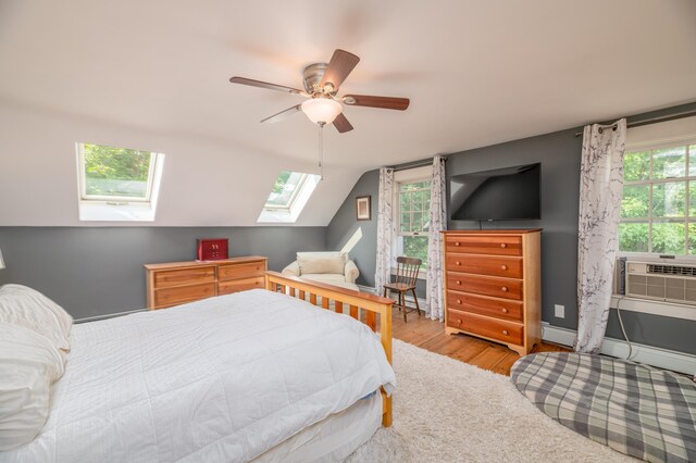 bedroom featuring ceiling fan, lofted ceiling with skylight, a baseboard heating unit, light hardwood / wood-style flooring, and cooling unit