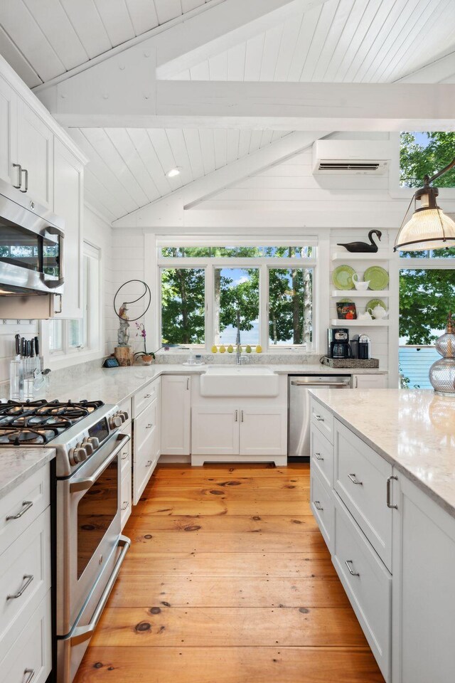 kitchen with light wood-type flooring, a wall mounted AC, lofted ceiling with beams, appliances with stainless steel finishes, and white cabinets