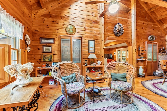 sitting room with wooden walls, light wood-type flooring, high vaulted ceiling, ceiling fan, and beam ceiling