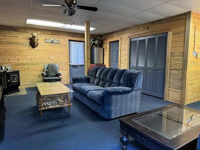 carpeted living room featuring a wood stove, a paneled ceiling, ceiling fan, and wooden walls