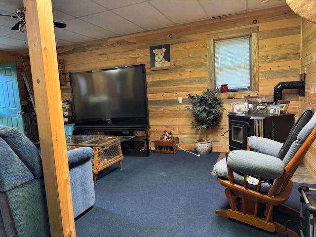 living room featuring wooden walls, a wood stove, a drop ceiling, carpet floors, and ceiling fan