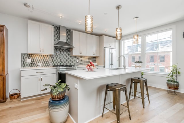 kitchen featuring appliances with stainless steel finishes, light hardwood / wood-style floors, an island with sink, white cabinets, and wall chimney range hood