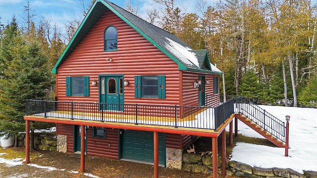 snow covered back of property featuring roof with shingles, an attached garage, a deck, stone siding, and log veneer siding