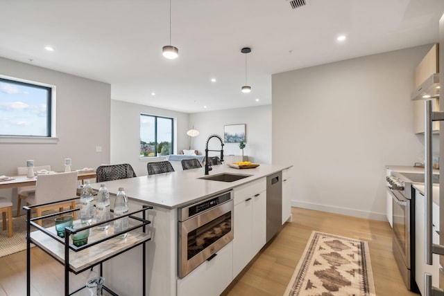kitchen with appliances with stainless steel finishes, white cabinetry, an island with sink, a sink, and light wood-style floors