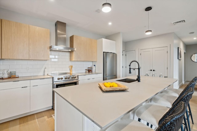 kitchen with appliances with stainless steel finishes, hanging light fixtures, wall chimney range hood, and an island with sink