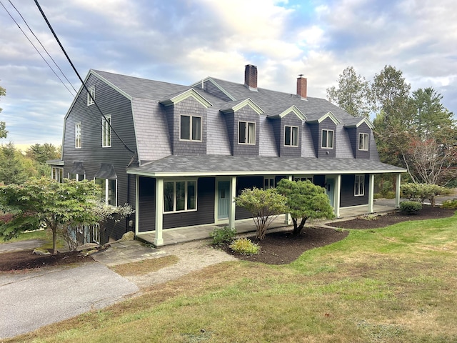 view of front of house with a gambrel roof, covered porch, a front yard, a chimney, and a shingled roof