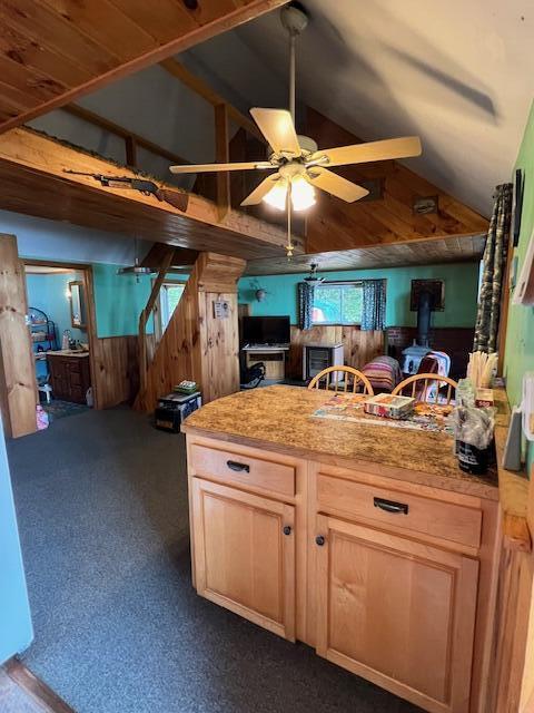 kitchen featuring a wood stove, a ceiling fan, lofted ceiling, wooden walls, and dark colored carpet